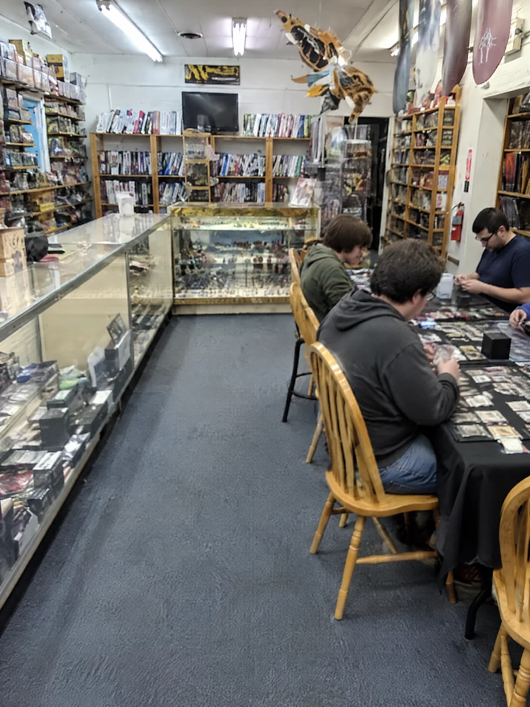 Inside Flights 2: glass cases of cards in front, wooden shelves of games, and players at folding tables under fluorescent lights.